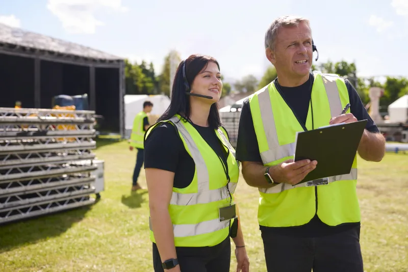 Male And Female Production Team With Headsets Setting Up Outdoor Stage For Music Festival Or Concert